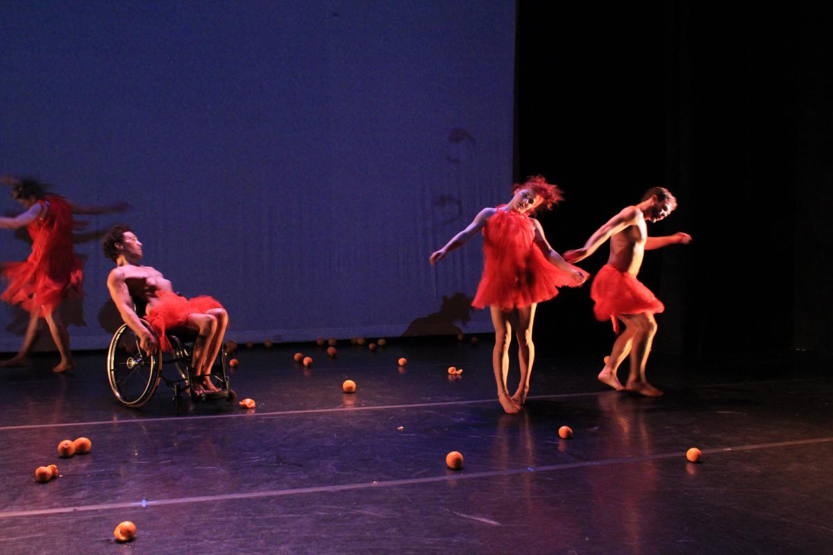 Oranges litter the stage. Four dancers in bright red costumes in synchronized motion; swirling, delirious, in an intoxicated state, moving across the stage. 
