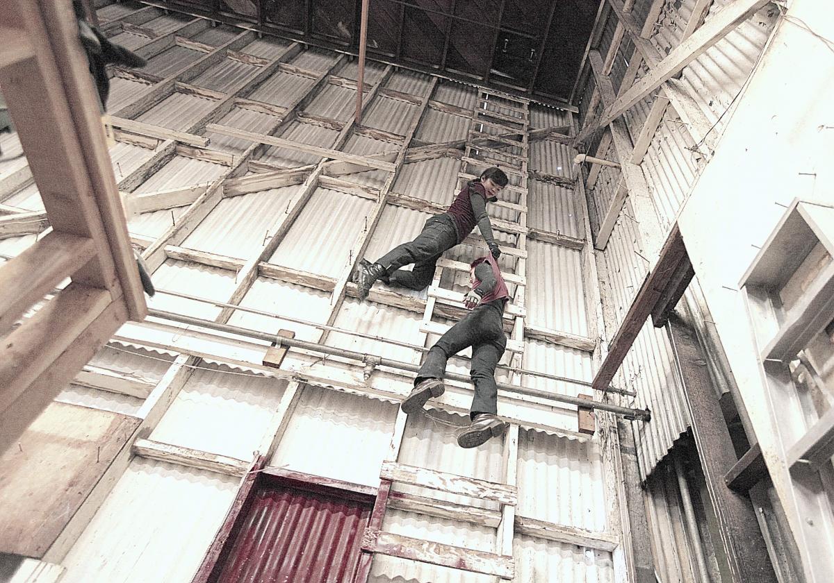 Megan Lowe and Sonsherée are high up on a white ladder in an industrial warehouse. Megan is holding onto the back of Sonsheree's shirt. Together they create an illusion that Sonsherée is dangling from the ladder and Megan is pulling her up by her shirt. 