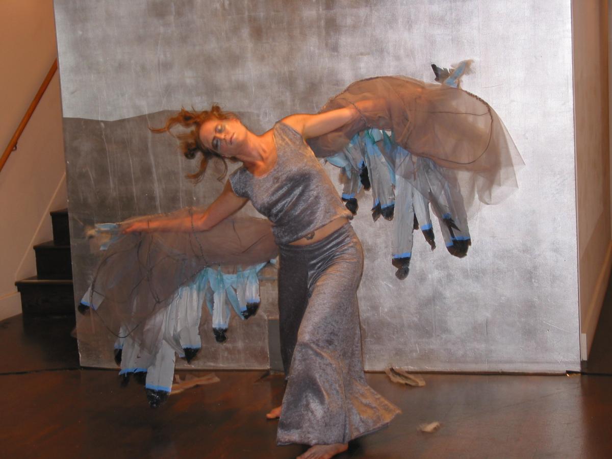 Dancer Amy Lewis, is wearing silver pants and a silver top. She has large feathered grey and silver wings strapped to her arms resembling a pigeon. She is in a deep lunge with her left leg forward and her head tipped to the right. Both wings are extended out parallel to the floor. She is caught in a moment between perching and taking flight.